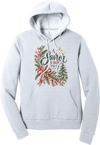 A Savior Is Born To Us hoodie in Ash, featuring watercolor evergreen and holly with a red ribbon bow, the words A Savior is born to us, and Isaiah 9:6