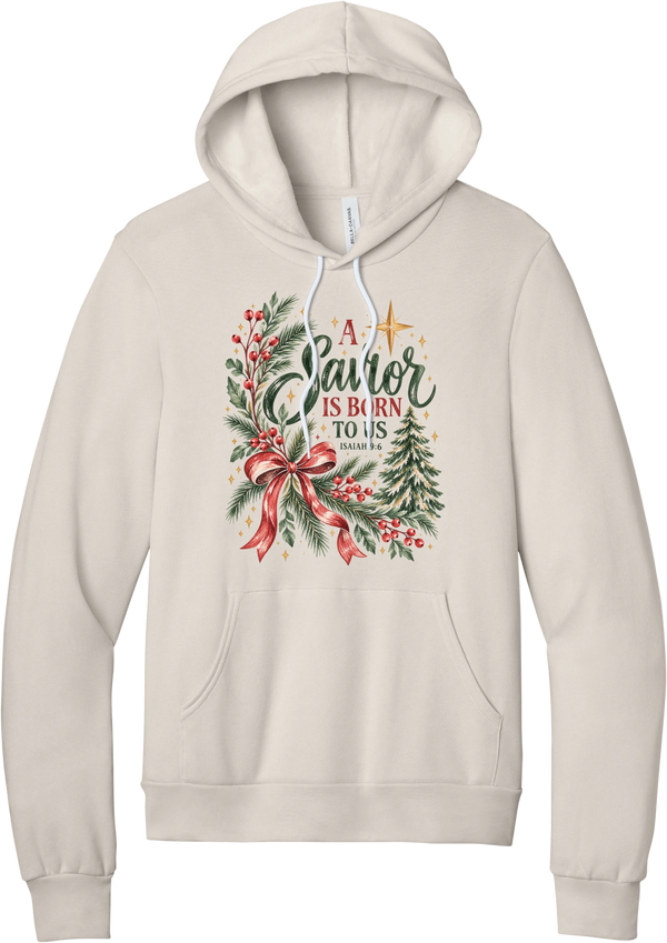 A Savior Is Born To Us hoodie in Heather Dust, featuring watercolor evergreen and holly with a red ribbon bow, the words A Savior is born to us, and Isaiah 9:6