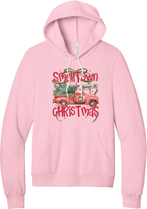Personalized Small Town Christmas hoodie in Pink, featuring Santa in a vintage red pickup truck hauling a Christmas tree past a personalized town road sign, with red Small Town Christmas lettering
