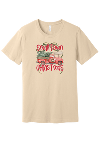 Personalized Small Town Christmas tee in Soft Cream, featuring Santa in a vintage red pickup hauling a Christmas tree past a road sign personalized with your town